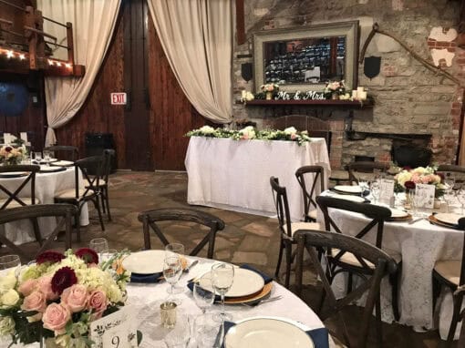 An indoor wedding reception setup with round tables, each with floral centerpieces and place settings. The head table has a Mr. and Mrs. sign.