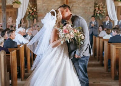 Bride and groom share a kiss inside a rustic chapel. Guests seated in wood pews look on. Draped white fabric and flowers decorate the space.