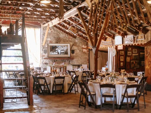 A warmly lit rustic barn is set up for an event with round tables covered in white tablecloths, each with place settings and centerpieces.