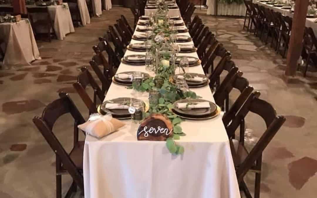 An event table setup with white linens, greenery centerpieces, and dark wood chairs in a rustic venue with wooden walls and a fireplace.