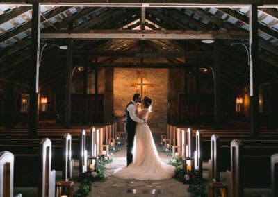 A bride and groom stand closely together in the aisle of a rustic chapel, with soft lighting, and a cross visible in the background.