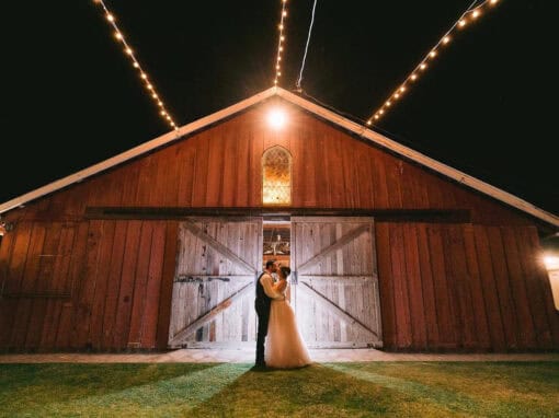 A bride and groom stand facing each other, under string lights in front of a large wooden barn at night.