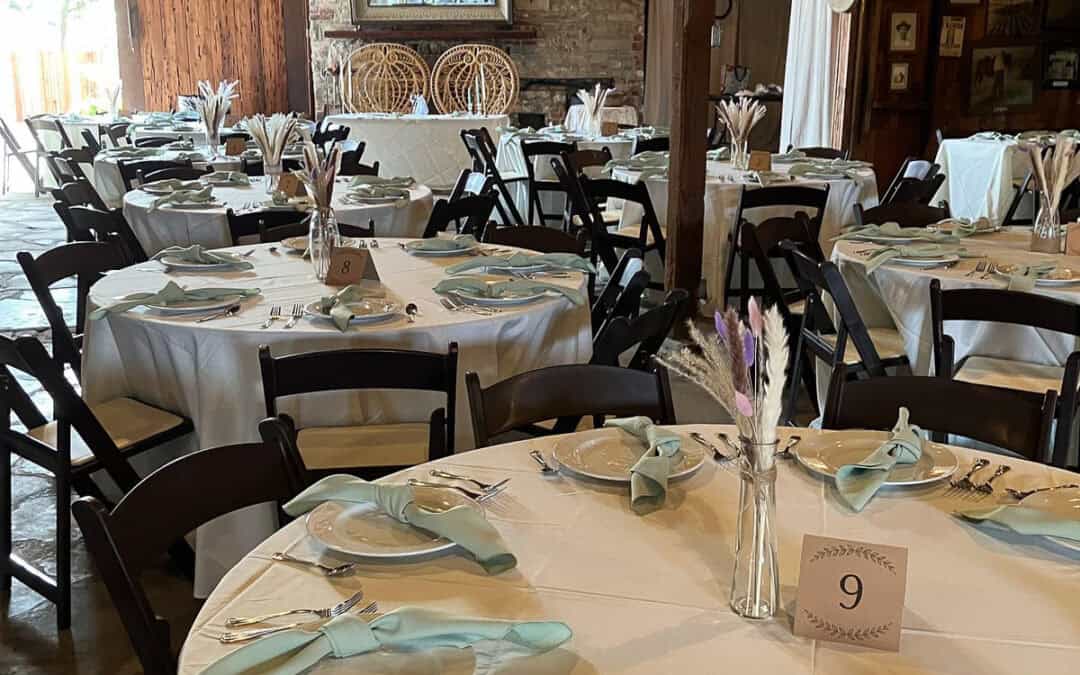 A rustic venue with round tables, adorned with white tablecloths, light green napkins, and floral centerpieces with string lights above.