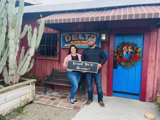 A couple stands in front of a red building holding a sign that reads, We Said Yes to the Venue! The background includes a DELTA sign.