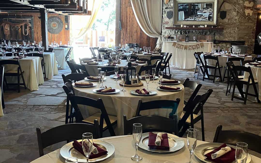 A rustic barn with round dining tables set with white tablecloths, maroon napkins, and glassware, arranged for a formal event.