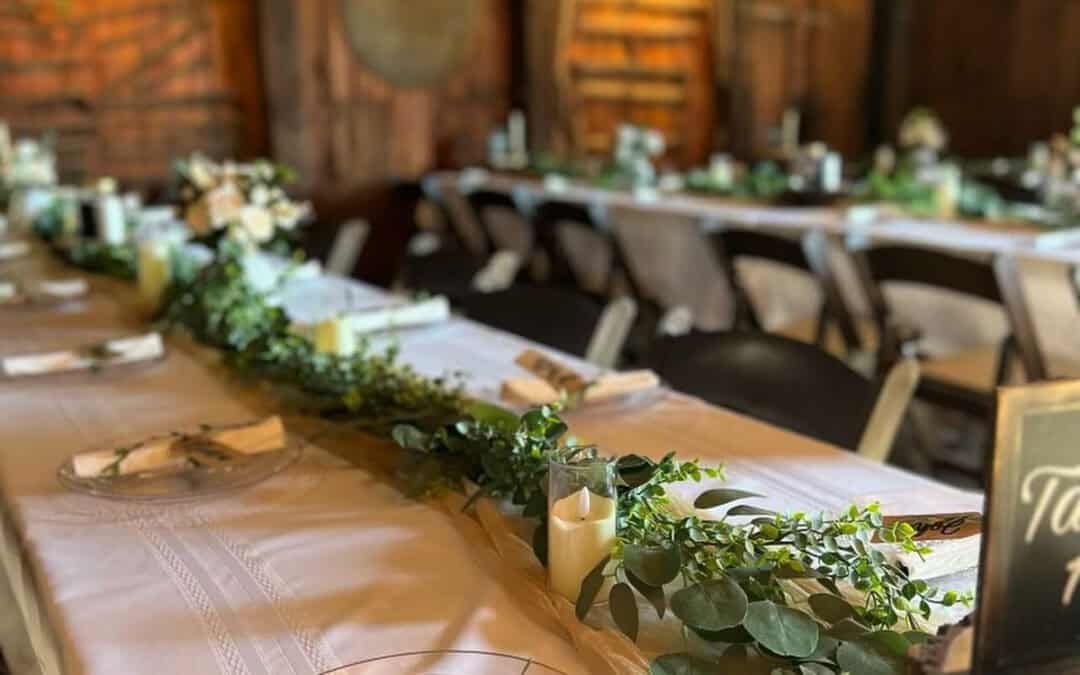 A rustic wooden hall setup with long banquet tables adorned with white tablecloths, greenery garlands, plates, and napkins.