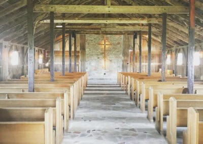 Interior of an empty chapel with wooden pews, a stone floor, exposed wooden beams, and a cross illuminated on the back wall.