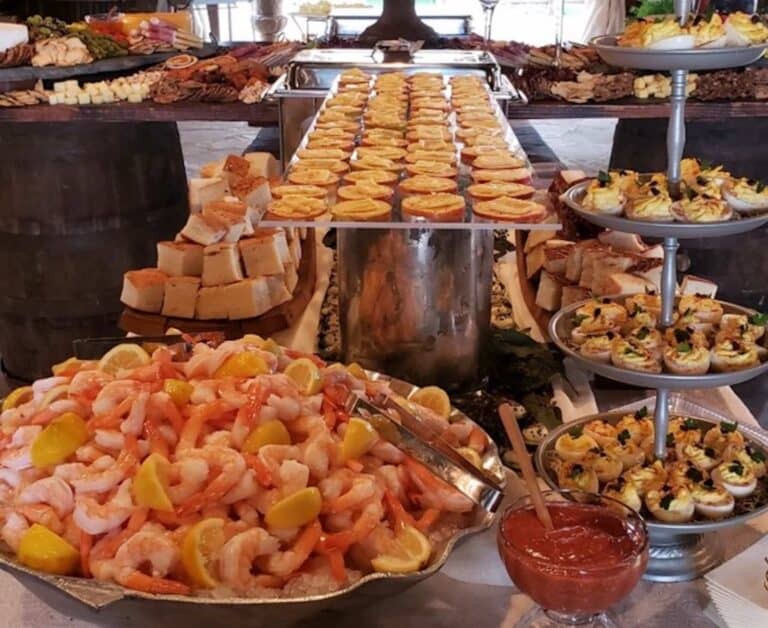 A buffet table featuring shrimp with lemon wedges, various tarts and pastries, bread pieces, and assorted appetizers.