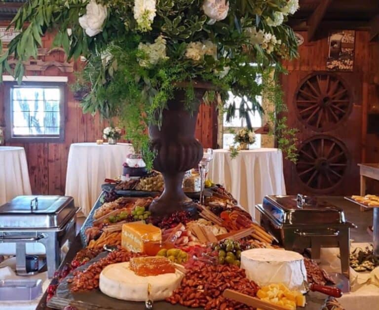 A large wood table covered with assorted cheeses, nuts, fruits, and a large floral arrangement is set up in a rustic venue.