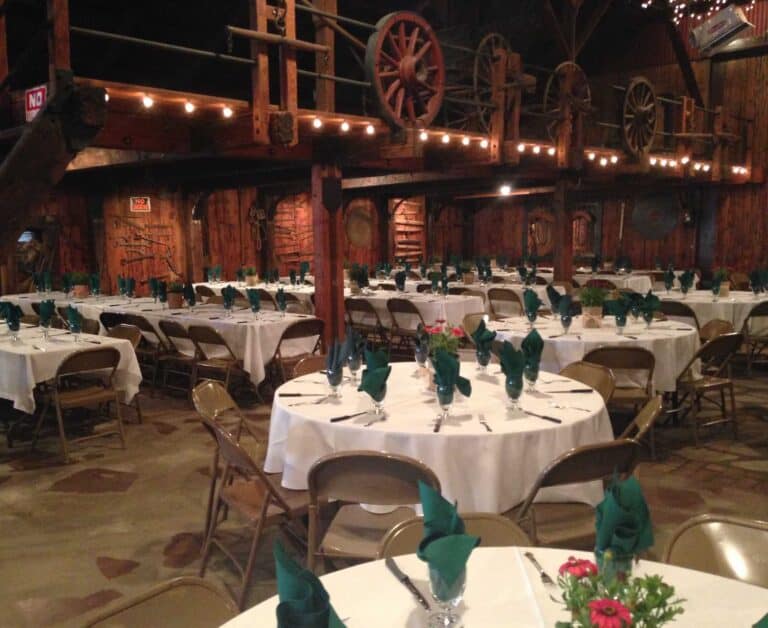 A rustic banquet hall with round tables in white tablecloths, set with green napkins. String lights are overhead.