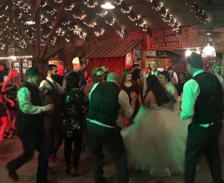 People in formal attire dancing under string lights in a rustic barn setting with a sign reading Delta Farms Party Barn in the background.