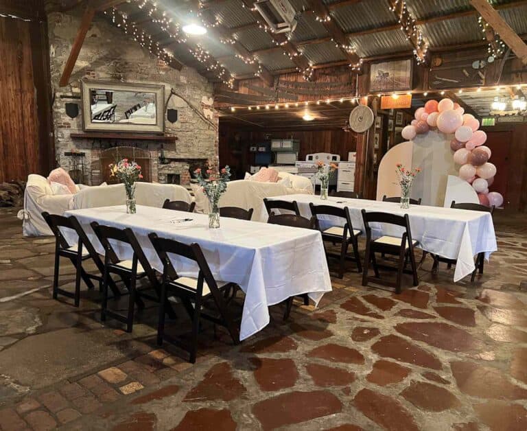 A rustic indoor venue with a stone floor and wood beams features two tables with white tablecloths, vase centerpieces, and pink balloons.