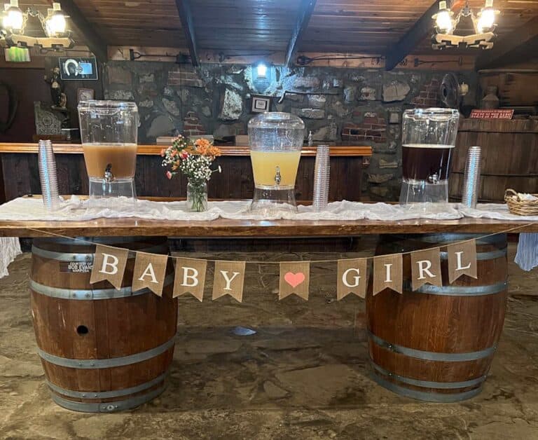 A rustic setup with 3 beverage dispensers on a wooden table adorned with a Baby Girl banner. The backdrop includes a stone wall and wood beams.