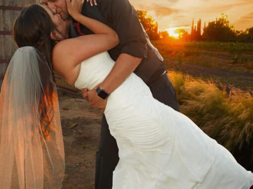 A bride and groom share a kiss at sunset, with the groom dipping the bride backward. The sky is vibrant with orange and pink hues.