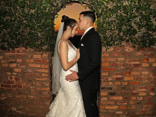 A groom kisses his bride's forehead under an arch of greenery with a brick wall and sunset in the background.