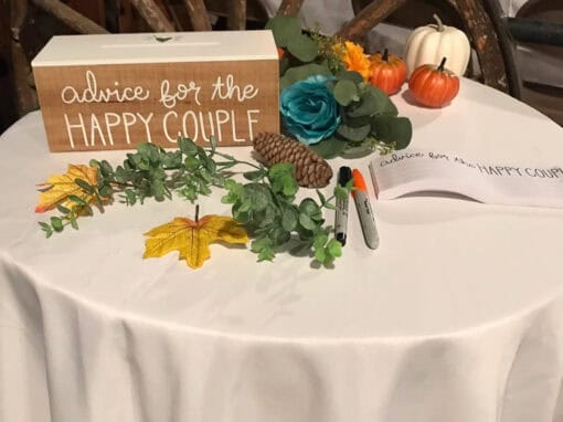 A table features a wood box reading Advice for the Happy Couple, pens, greenery, a blue rose, small pumpkins, and 2 yellow leaves.