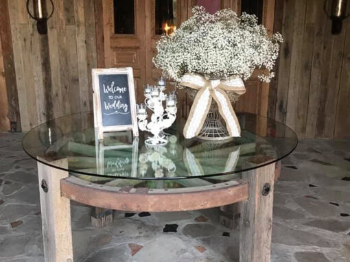 A round wood table with a glass top holds a sign that says Welcome to our Wedding, a bouquet of flowers, and decorative candles.