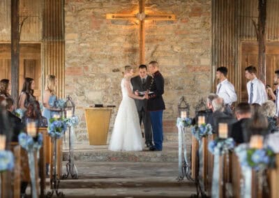 A bride and groom stand at the altar during a wedding ceremony in a rustic chapel, surrounded by bridesmaids, groomsmen, and guests.