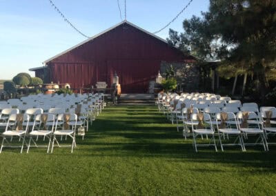 An outdoor wedding setup with rows of white folding chairs, a red barn, and string lights overhead.