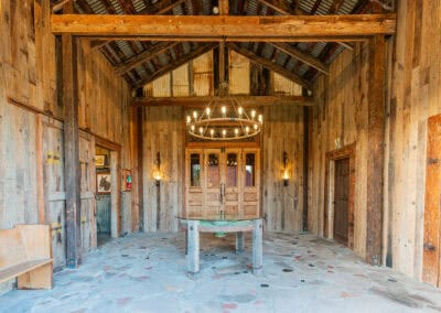 A rustic room with wood walls and beams, a central table, chandelier, and stone floors. 2 open wooden doors reveal a chapel beyond.