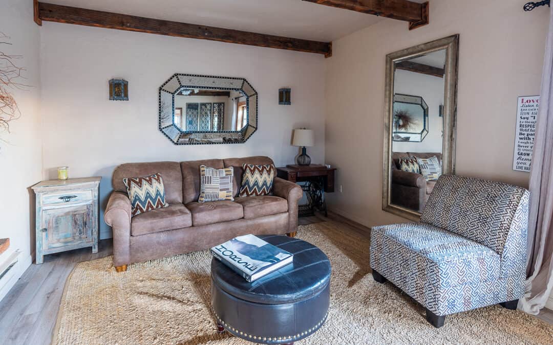 A room with a brown sofa, a single chair, a round black ottoman, a beige rug, two wall mirrors, and a small side table with a lamp.