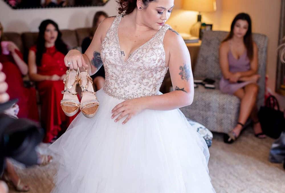 A bride in a white wedding dress holds a pair of gold shoes in her hand, standing indoors with several seated women in the background.